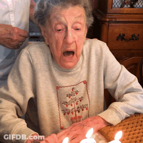 An Elderly Woman Sitting At A Table With Candles In Her Hand GIF