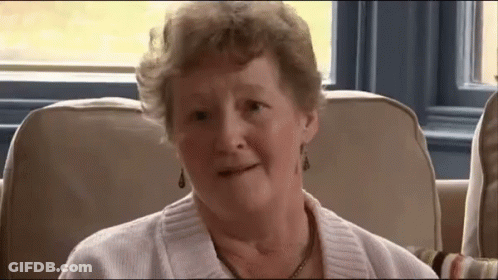 An Elderly Woman Wearing A White Sweater And Earrings Is Sitting On A Couch GIF