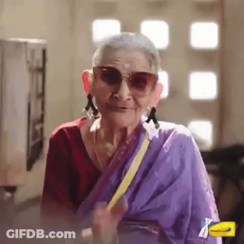 An Elderly Woman Wearing Sunglasses And A Purple Sari Is Smiling And Waving . GIF