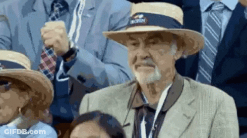 An Older Man Wearing A Straw Hat With A Nyc Logo On It Is Sitting In A Crowd Of People . GIF