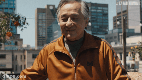 An Older Man Wearing An Orange Jacket With The Hashtag #Karatekidmovie On The Bottom GIF