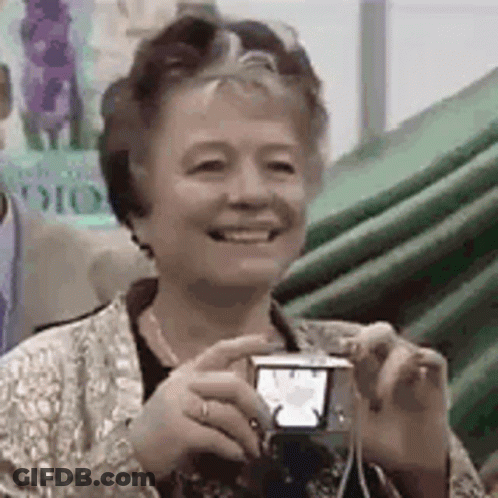 An Older Woman Is Smiling While Holding A Small Camera . GIF