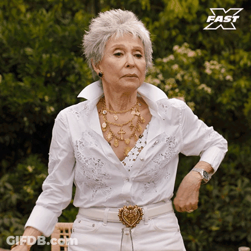 An Older Woman Wearing A White Shirt And White Pants Is Standing In Front Of A Sign That Says Fast X GIF