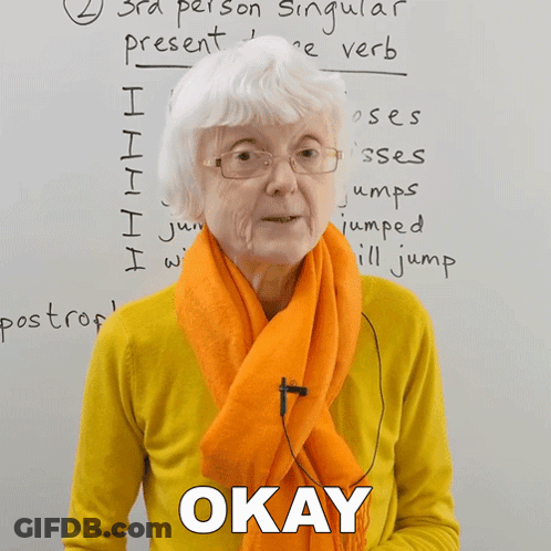 An Older Woman Wearing Glasses And An Orange Scarf Says Okay In Front Of A Whiteboard GIF