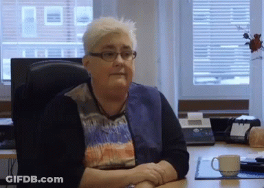 An Older Woman Wearing Glasses Sits At A Desk With Her Hands Folded GIF