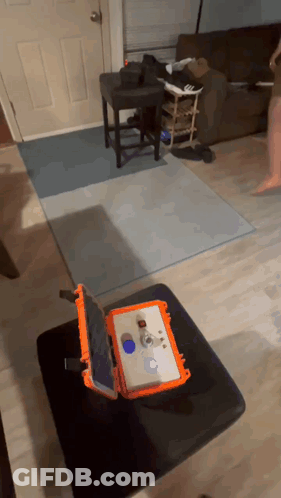 An Orange Case With A Tablet Inside Of It Sits On A Black Ottoman In A Living Room GIF