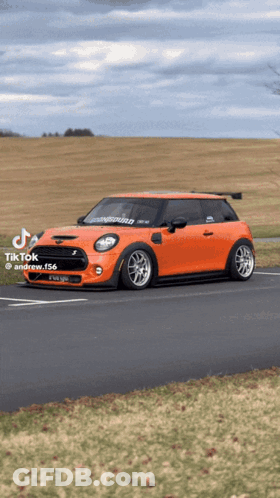 An Orange Mini Cooper Is Parked On The Side Of The Road With A Field In The Background GIF