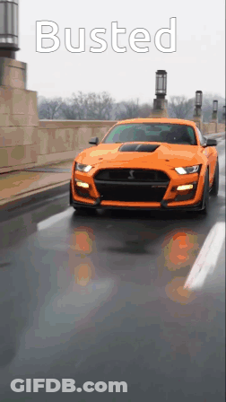 An Orange Mustang Is Driving Down A Street With The Word Busted On The Bottom GIF