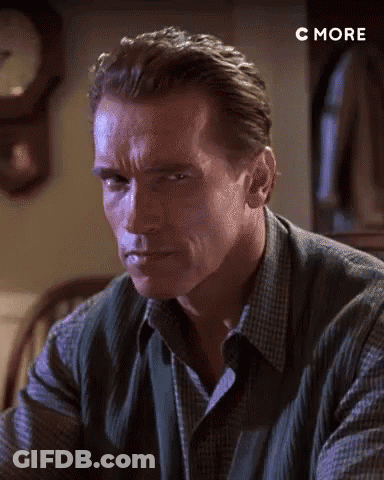 Arnold Schwarzenegger Is Sitting At A Table In Front Of A Clock And Looking At The Camera . GIF