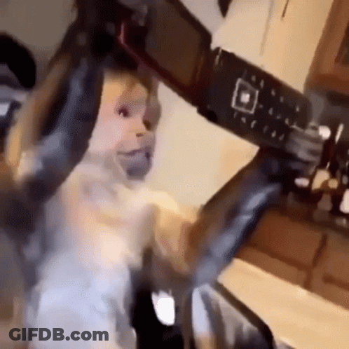 Baby Holding Camera Making Funny Face In Kitchen GIF