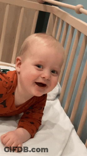 Baby In Orange Shirt Laying In Wooden Crib GIF