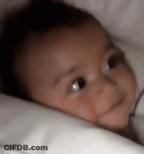 Baby Laying On Bed With White Blanket Looking At Camera GIF