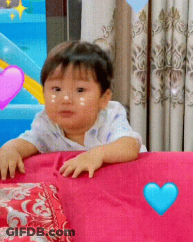 Baby Sitting On Bed With Pink Pillow And Blue Heart GIF