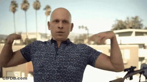 Bald Man In Blue Shirt Flexing Muscles Holding Gun GIF