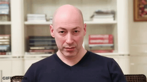 Bald Man In Blue Shirt Sitting In Front Of Bookshelf GIF