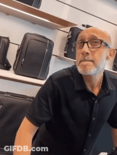 Bald Man In Glasses And Black Shirt In Front Of Suitcases GIF