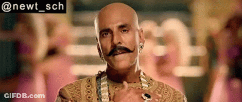 Bald Man In Gold Dress With Mustache, Earrings, And Necklace GIF