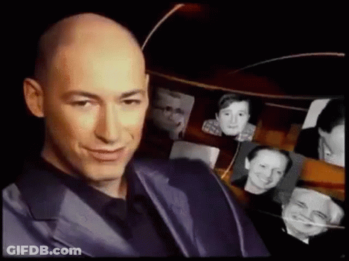 Bald Man In Suit Smiling In Front Of Pictures GIF