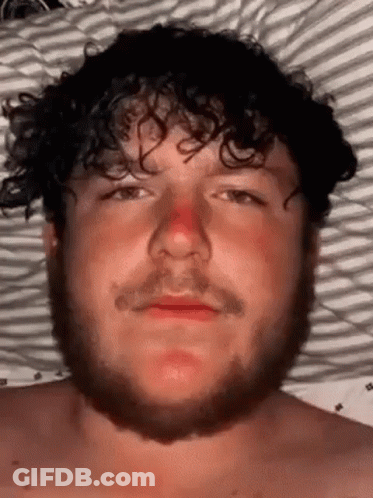 Bearded Curly-Haired Man Laying In Bed With Red Nose GIF