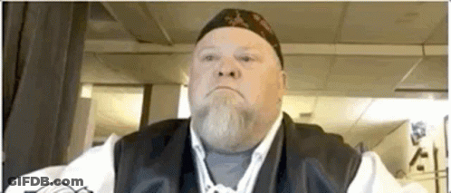 Bearded Man In Hat And Vest Looks At Camera GIF