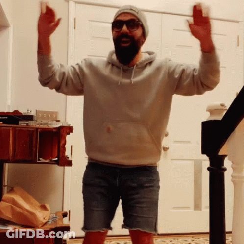 Bearded Man In Shorts And Hoodie Standing In Front Of Door With Arms In Air GIF