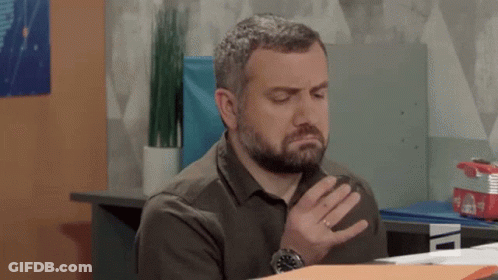 Bearded Man Sitting At Desk With Hand To Chest GIF
