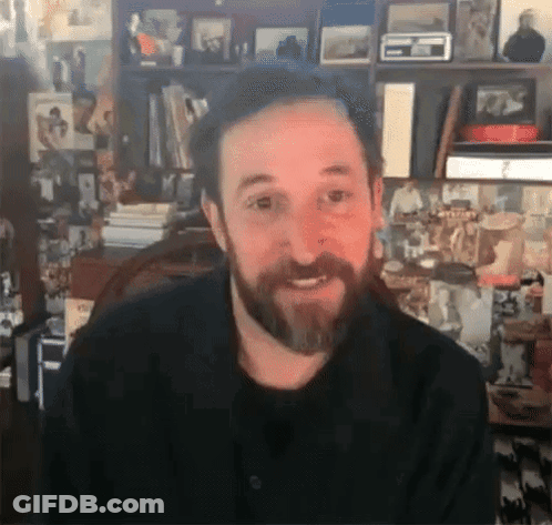 Bearded Man Smiling In Front Of Bookshelf With Pictures GIF