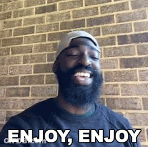 Bearded Man Smiling In Front Of Brick Wall Saying Enjoy Enjoy GIF