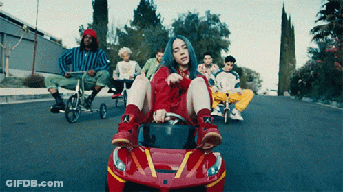 Billie Eilish Sitting In Red Toy Car With Group Riding Bicycles Behind GIF