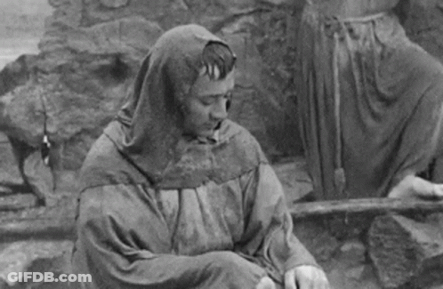 Black And White Man In Hooded Cloak Sitting On Ground GIF