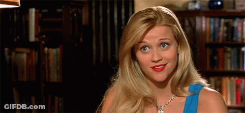 Blonde Woman In Blue Top Smiling In Front Of Bookshelf GIF