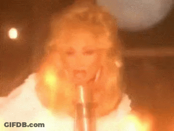 Blonde Woman Singing Into Microphone In Front Of Fire GIF