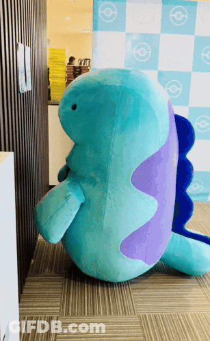 Blue And Purple Stuffed Animal Sitting In Front Of Checkered Wall GIF