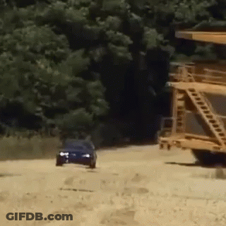 Blue Car Driving Down Dirt Road Past Yellow Truck GIF