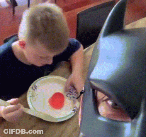Boy Eating Plate Of Food Next To Batman Mask GIF