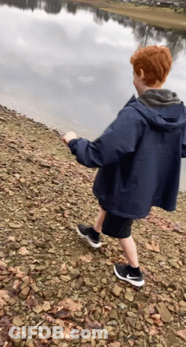 Boy In Blue Jacket Walking On Rocky Shoreline GIF