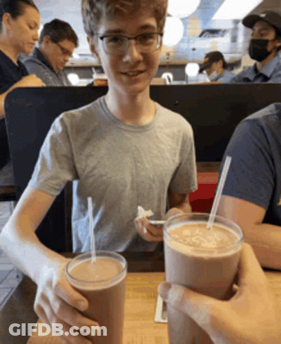 Boy With Glasses Sitting At Table With Two Milkshakes GIF