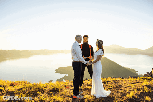 Bride And Groom Holding Hands On Hill Overlooking Lake GIF