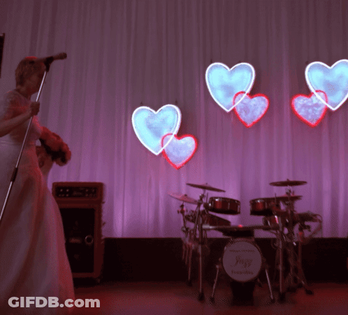 Bride Singing Into Microphone In Front Of Drum Set With Hearts GIF