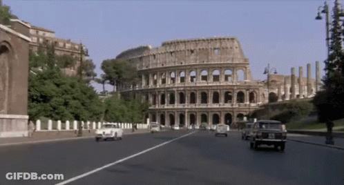 Cars Are Driving Down A Street In Front Of A Large Building Called The Colosseum GIF