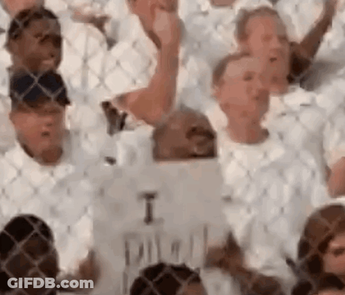 Crowd In White Shirts Holding Signs Behind Chain Link Fence GIF