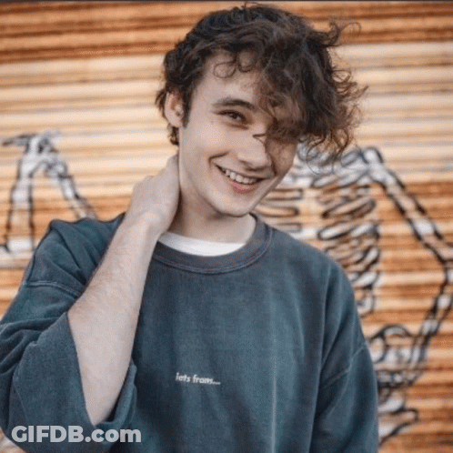 Curly Haired Man Smiling Next To Skeleton Wall GIF