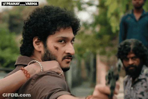 Curly Haired Man With Beard Holding Gun In Front Of Pranay Varma Sign GIF