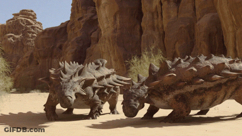 Dinosaurs Standing In Sand Near Rock Formation GIF