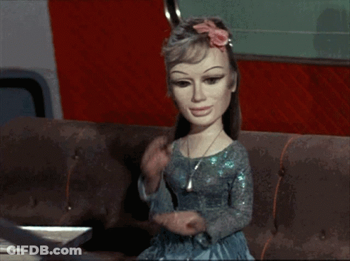 Doll With Flower In Hair Sitting On Brown Couch GIF