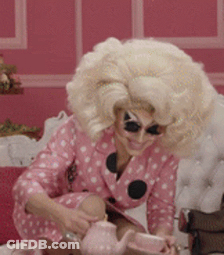Drag Queen With Blonde Wig And Teapot On Bed GIF