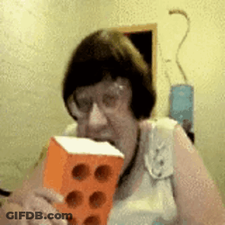 Elderly Woman Holding Brick In Mouth With Glasses GIF