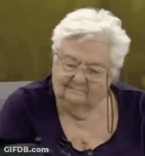 Elderly Woman In Glasses And Purple Shirt Smiling On Couch GIF
