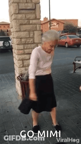 Elderly Woman Next To Brick Wall Holding Hat GIF