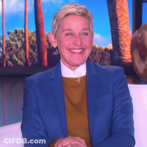 Ellen Degeneres Is Wearing A Blue Suit And A Yellow Sweater And Smiling While Sitting On A Couch . GIF